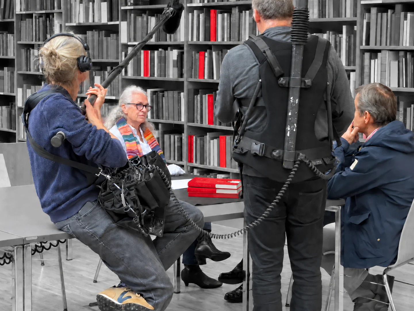Gesprächssituation mit Aufnahmeteam vor Bücherregalen. Personen sind farbig dargestellt, einzelne Bücher leuchten rot.