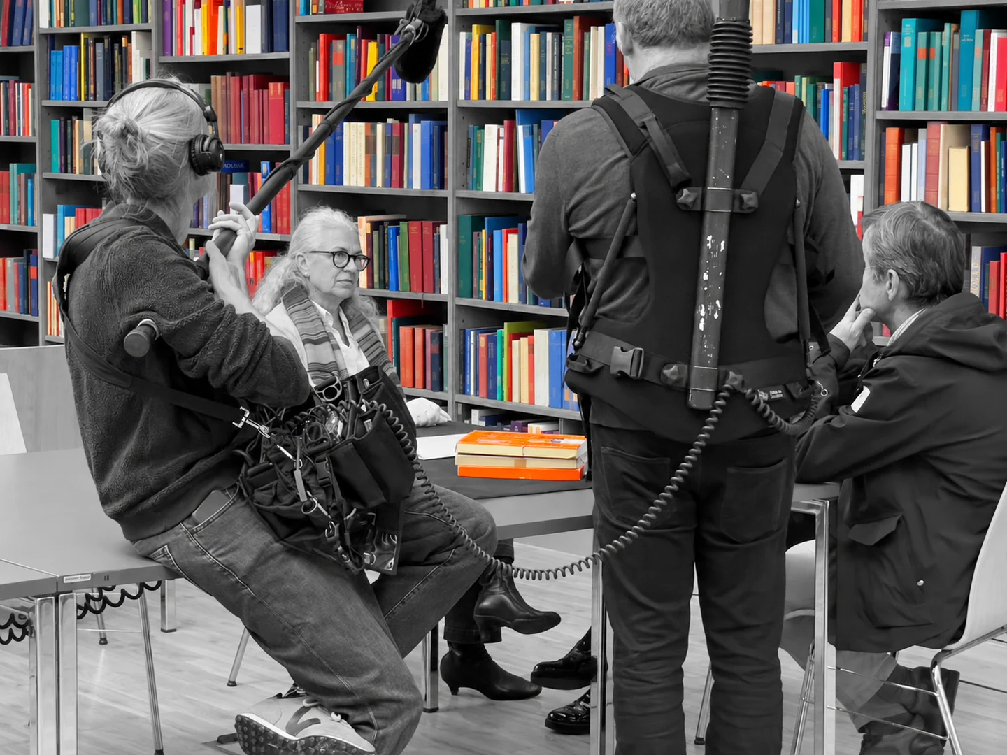 Gesprächssituation mit Aufnahmeteam vor Bücherregalen. Die Bücher sind farbig hervorgehoben, die Personen schwarzweiß dargestellt.