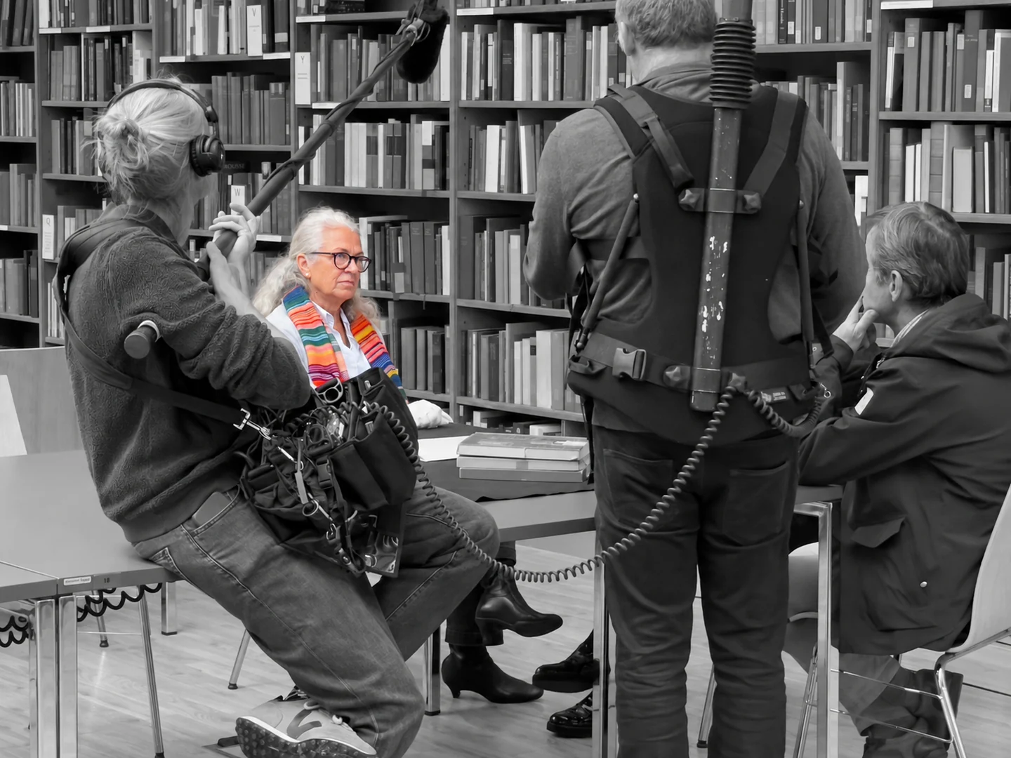Gesprächssituation mit Aufnahmeteam vor Bücherregalen. Eine sitzende Gesprächspartnerin ist farbig hervorgehoben, ihre Umgebung ist schwarzweiß.