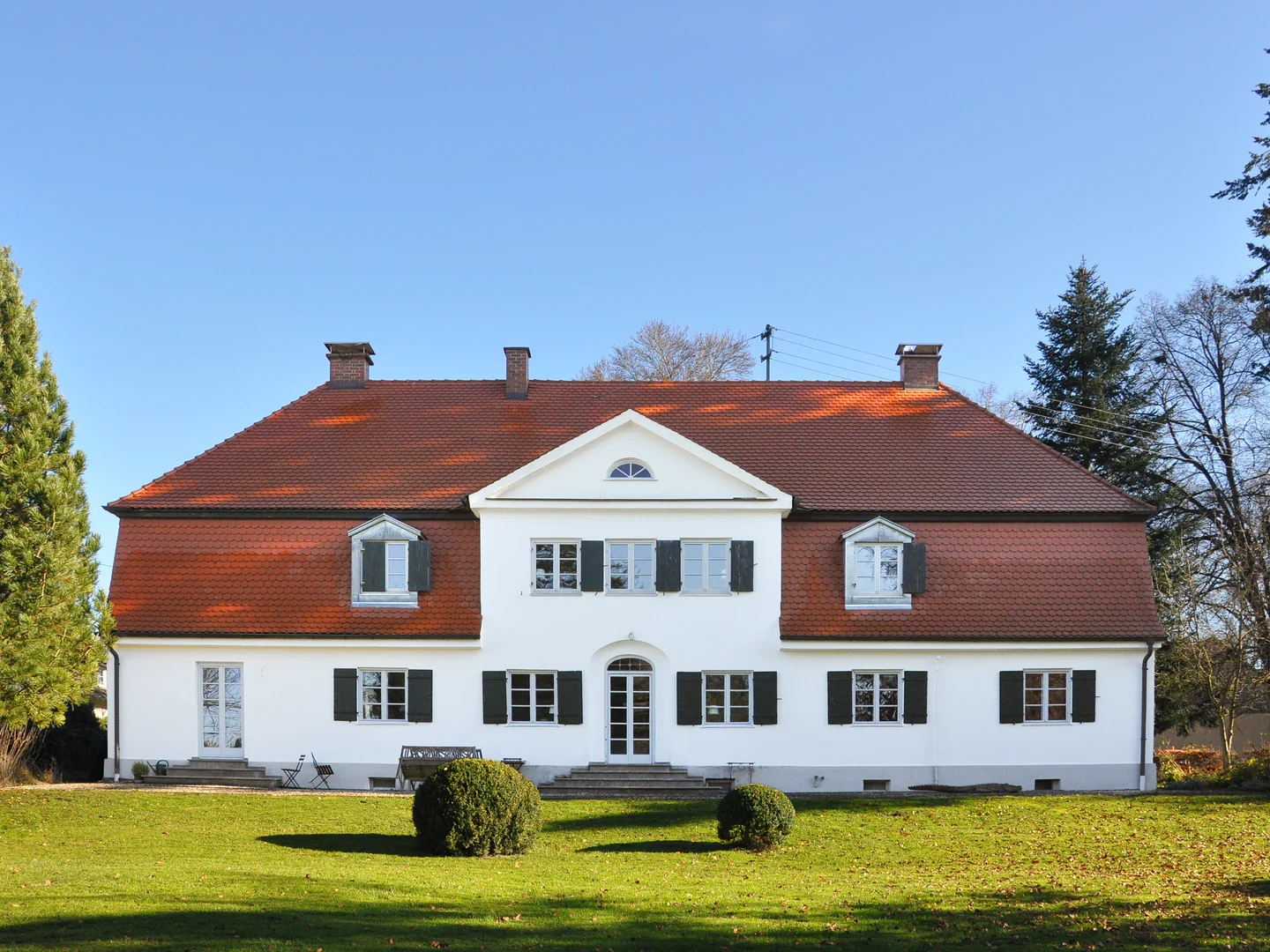Frontalansicht eines weißen Gebäudes mit rotem Dach, Fensterläden und Garten.